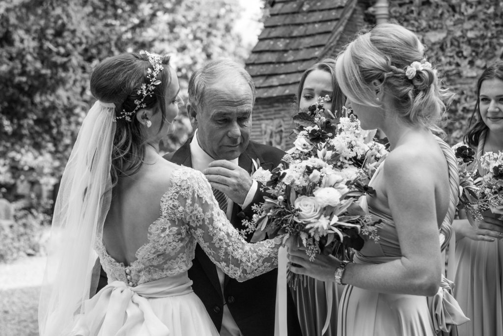 Bride and her father with bridesmaids ready to go into church. Hair is half up with relaxed curls. A hair vine crowning the style and cathedral length veil in the back