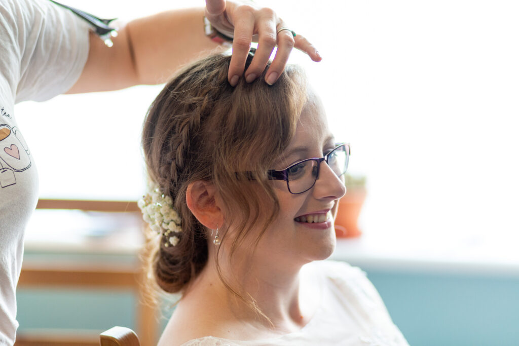 A hand going through the fringe section of a beautiful bridal hair up separating to create texture. A hairstylist doing the finishing touches to a smiling bride wearing glasses. Happy times.